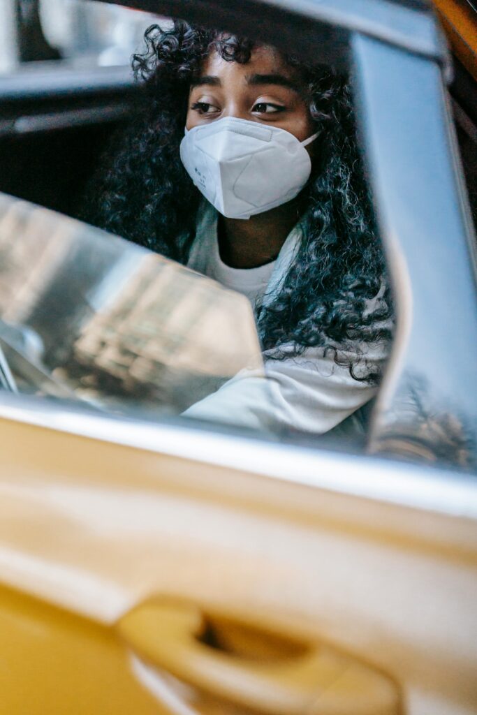 A woman with curly hair wearing a mask sits in a taxi, gazing out the window, reflecting the urban streets.